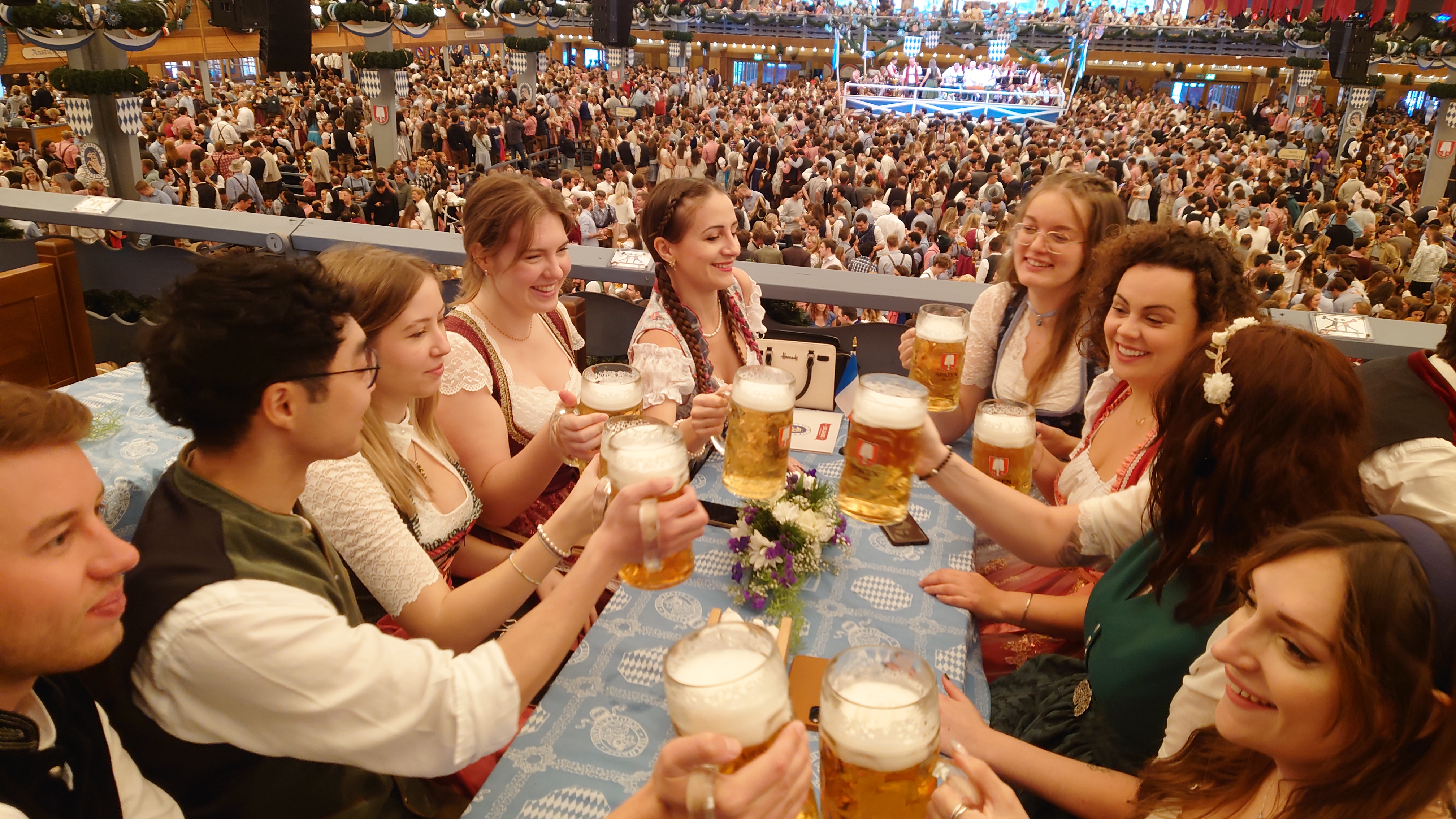 Oktoberfest i München 2022 - ungdomar i folkdräkt, skålar med ölkrus vid ett bord i öltältet.